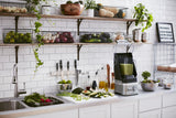 A modern kitchen with white tiled walls and a white countertop. A KitchenAid Commercial Blender filled with green smoothie sits beside fresh vegetables, knives, and bottles. Open shelves above hold produce baskets, bowls, and kitchenware. A sink is visible on the left.