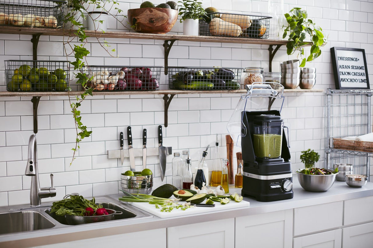 A modern kitchen with white tiles and counter, featuring a KitchenAid Commercial Blender with 60 oz. capacity filled with green smoothie, surrounded by chopped vegetables, salad bowl, oil bottles, and baskets of fresh produce on wall shelves.