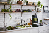 A modern kitchen with white tiles and counter, featuring a KitchenAid Commercial Blender with 60 oz. capacity filled with green smoothie, surrounded by chopped vegetables, salad bowl, oil bottles, and baskets of fresh produce on wall shelves.