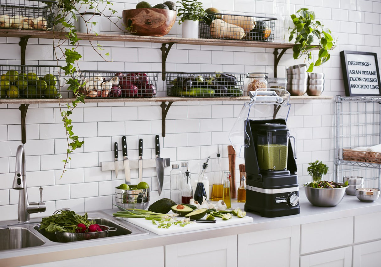 A bright kitchen with white tile walls, open shelves of fresh produce and plants, and a countertop featuring a KitchenAid Commercial Blender with 60 oz. capacity blending green smoothie, plus veggies, knives, bottles, and salad by the sink.