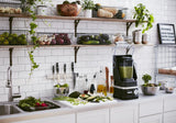 A bright kitchen with white tile walls, open shelves of fresh produce and plants, and a countertop featuring a KitchenAid Commercial Blender with 60 oz. capacity blending green smoothie, plus veggies, knives, bottles, and salad by the sink.