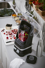 A KitchenAid Commercial Blender with a clear, open lid contains chopped red and green fruits on a kitchen counter. Nearby are stacked plastic cups, a cutting board with pomegranate and berries, and baskets of fresh fruit on a shelf.