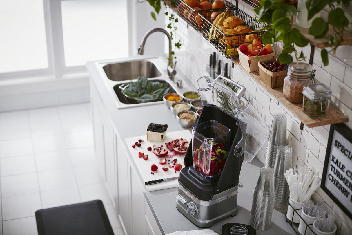 A modern kitchen counter with a BPA free blender filled with berries and greens. Surrounding it are chopped pomegranates, containers of fruits and vegetables, glasses, paper cups, a sink, and bright shelves stocked with plants and supplies.