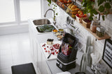 A modern kitchen countertop with a KitchenAid Commercial Blender containing pomegranate and berries, a cutting board with fruit and a knife, and bowls of ingredients near a sink. Shelves hold fresh produce and jars, with plants and cups along a white-tiled wall.
