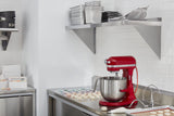 A modern kitchen with white tile walls and stainless steel counters. A KitchenAid Commercial Stand Mixer sits on the counter near rows of colorful macarons. Above are metal shelves with stacked pans, trays, and a colander.