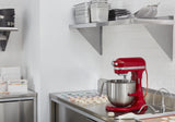 A shiny red KitchenAid Commercial Stand Mixer sits on a stainless steel countertop in a modern kitchen with white subway tile walls. Nearby, pastel macarons are arranged on parchment, while baking trays and bowls rest on open shelves above.