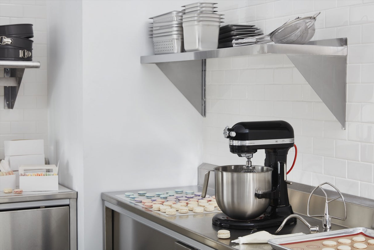 A modern kitchen features a black 8 quart stand mixer on the counter beside a sink, baking sheets lined with colorful macarons, stacked metal bakeware above, and white subway tile backsplash. Stainless steel surfaces create a clean, organized workspace.