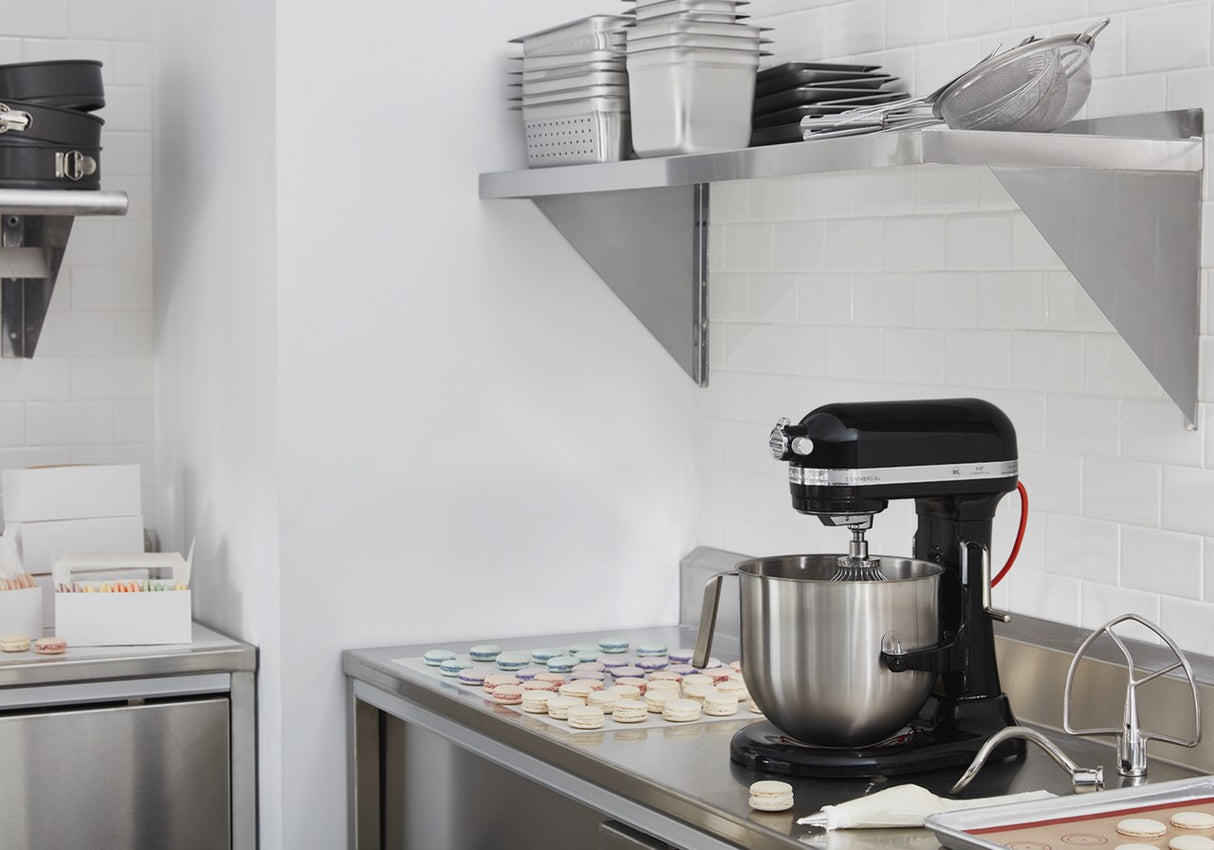 A stainless steel countertop holds a black KitchenAid Commercial Stand Mixer with a metal bowl, a piping bag, and rows of pastel-colored macarons. Shelves above display stacked trays and kitchenware. The kitchen is spotless and organized.