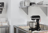 A stainless steel countertop holds a black KitchenAid Commercial Stand Mixer with a metal bowl, a piping bag, and rows of pastel-colored macarons. Shelves above display stacked trays and kitchenware. The kitchen is spotless and organized.