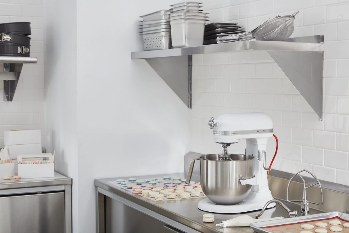 A clean, modern kitchen features a white KitchenAid Commercial Stand Mixer with an 8 quart bowl on a stainless steel counter. Macarons are arranged on trays nearby, beneath metal shelves stacked with pans and containers.
