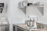A clean, modern kitchen features a white KitchenAid Commercial Stand Mixer with an 8 quart bowl on a stainless steel counter. Macarons are arranged on trays nearby, beneath metal shelves stacked with pans and containers.
