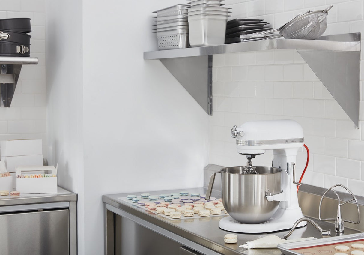 A clean, modern kitchen with white subway tile walls and stainless steel surfaces. A KitchenAid Commercial Stand Mixer with an 8 quart bowl is on the counter next to colorful macarons. Metal shelves above hold pans, bowls, and baking tools.