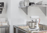 A clean, modern kitchen with white subway tile walls and stainless steel surfaces. A KitchenAid Commercial Stand Mixer with an 8 quart bowl is on the counter next to colorful macarons. Metal shelves above hold pans, bowls, and baking tools.