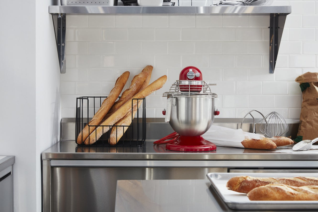 A modern kitchen with a metal counter holds a red KitchenAid Commercial Stand Mixer with an 8 quart bowl, a wire rack with baguettes, a metal whisk, a brown paper bag, and a baking tray of baguettes. White subway tiles and metal shelving are visible behind.