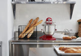 A modern kitchen counter with a KitchenAid Commercial Stand Mixer and 8 quart bowl, a metal tray of baguettes, a wire basket of baguettes, a whisk, and paper bag. The backdrop features white subway tile and a metal shelf with bowls and kitchen items.