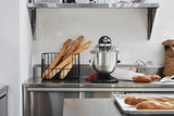 A modern kitchen counter features several baguettes in a wire basket, a KitchenAid Commercial Stand Mixer with an 8 quart bowl, a wire whisk, a brown paper bag, and fresh bread—all set against white subway tile and metal shelves.