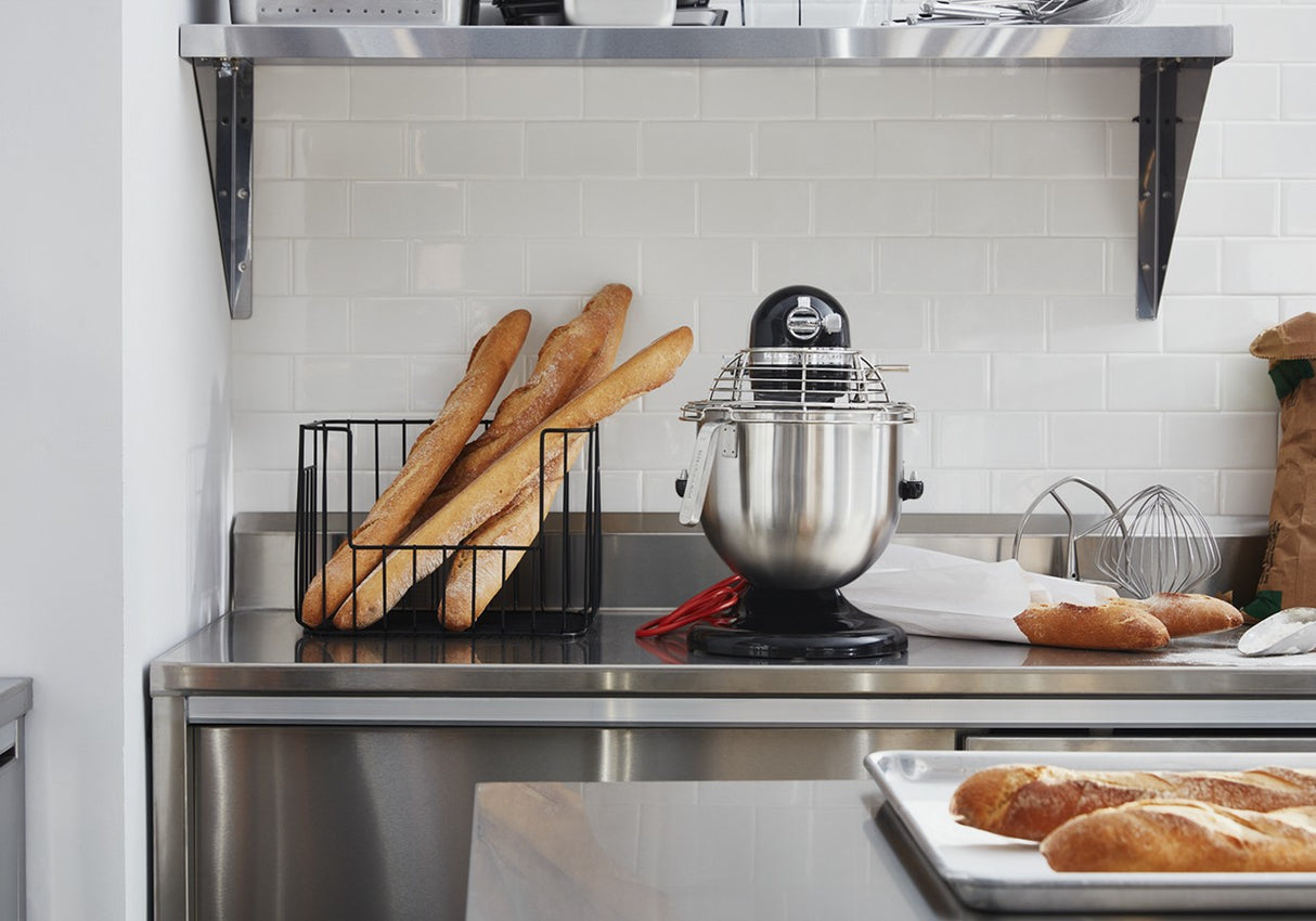 A stainless steel kitchen counter holds a metal bread rack with several baguettes, a KitchenAid Commercial Stand Mixer with an 8 quart bowl and wire guard, and a red spatula. Nearby, baguettes rest on a baking sheet; white subway tiles line the wall.