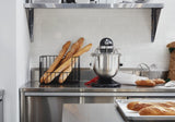 A stainless steel kitchen counter holds a metal bread rack with several baguettes, a KitchenAid Commercial Stand Mixer with an 8 quart bowl and wire guard, and a red spatula. Nearby, baguettes rest on a baking sheet; white subway tiles line the wall.