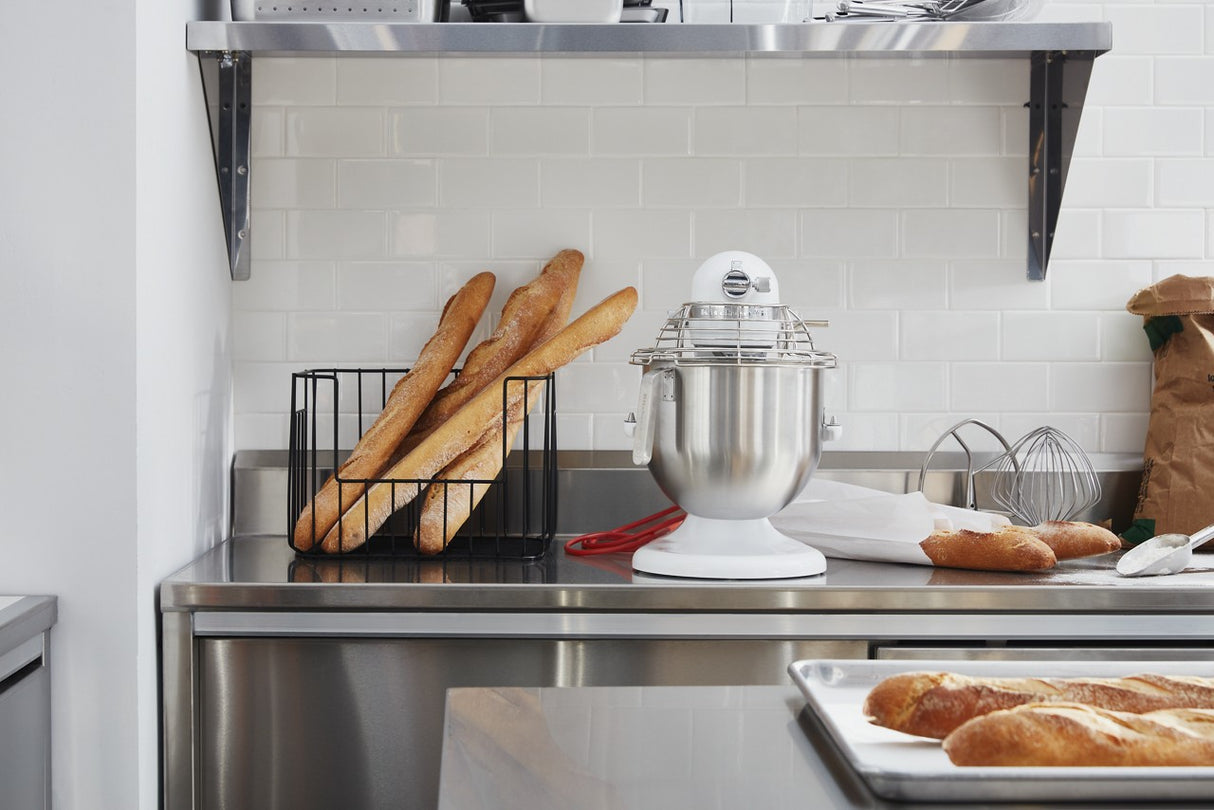 A clean, modern kitchen with a metal countertop features a KitchenAid Commercial Stand Mixer with an 8 quart bowl, a basket of baguettes, wire whisk, brown paper bag, tray of baked bread, white subway tile backsplash, and a metal shelf in the background.