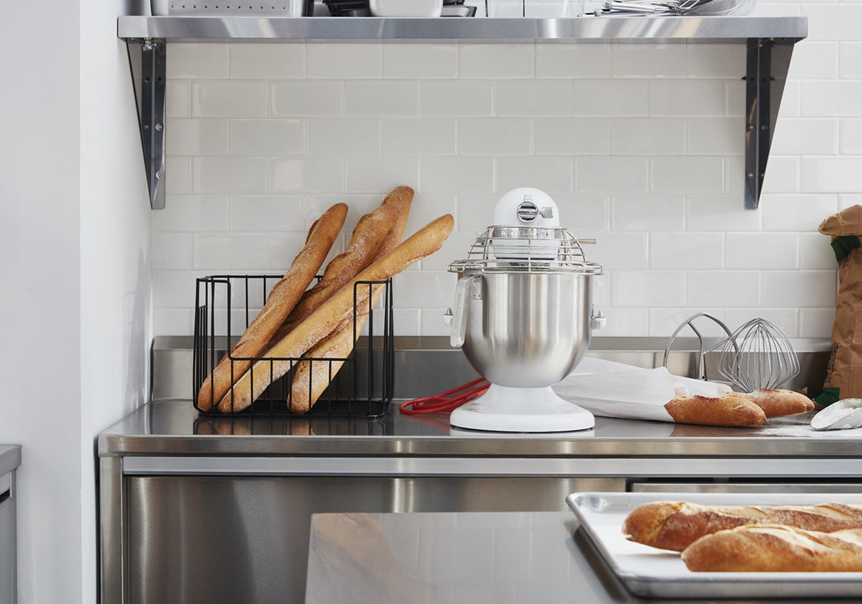 A modern kitchen with a metal countertop displays baguettes in a black wire basket, an 8 quart bowl KitchenAid Commercial Stand Mixer, a wire whisk, loaves of bread, and a baking tray. White subway tiles and a metal shelf are visible in the background.