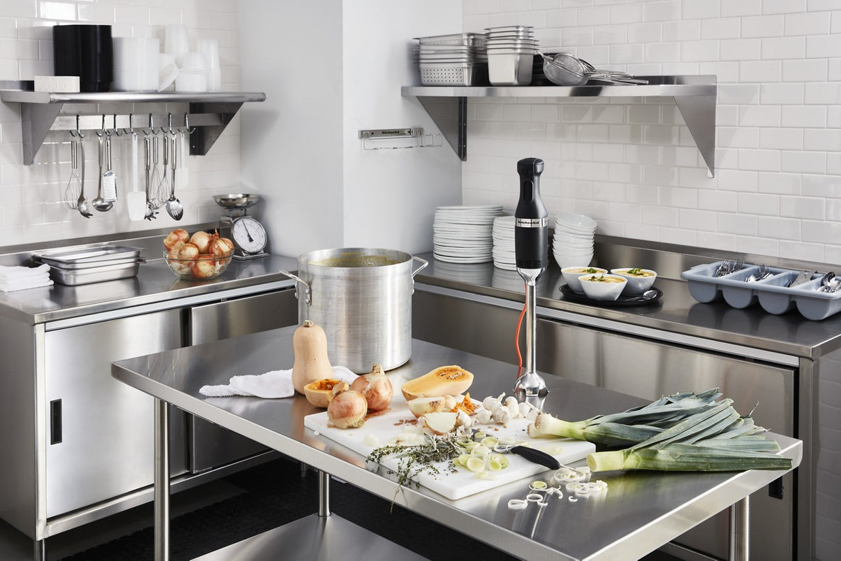 A commercial kitchen with stainless steel counters, shelves, and appliances. Vegetables, herbs, and a KitchenAid commercial immersion blender with a 12 stainless steel blending arm are on the prep table amid neatly arranged bowls, plates, and utensils.