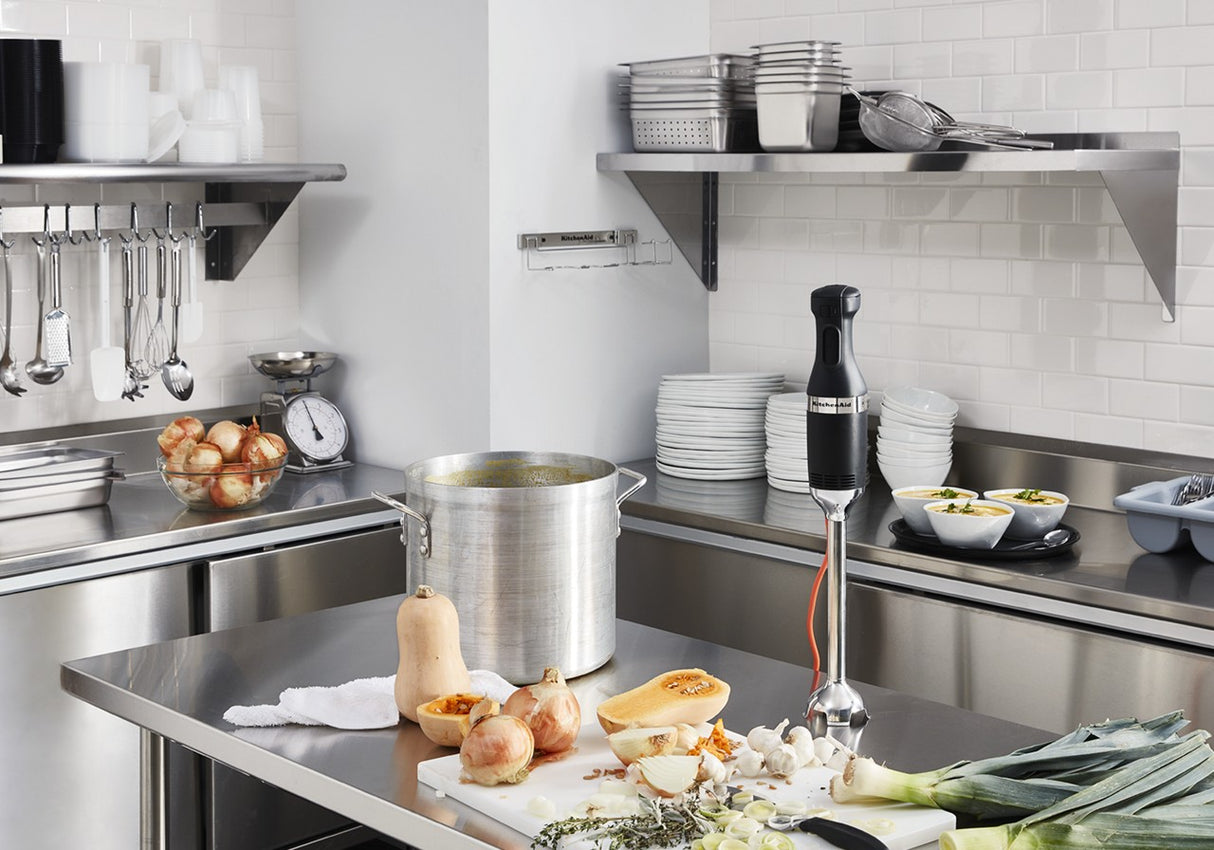 A commercial kitchen with stainless steel counters holds a large pot of soup, a heavy duty immersion blender, squash, onions, and leeks on a cutting board. Shelves display stacked plates, bowls, colanders, utensils, and apples. The scene is clean and organized.