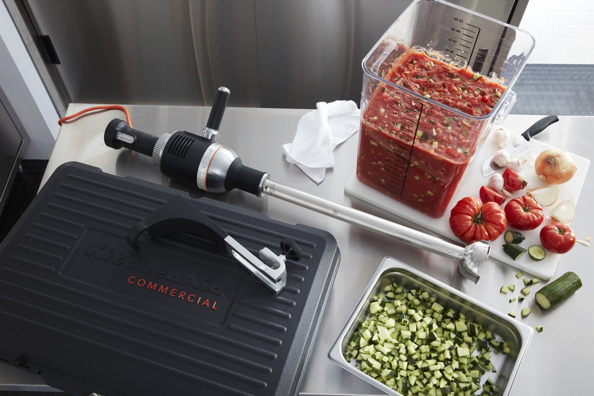 A commercial kitchen counter displays a heavy duty immersion blender, a black KitchenAid case, chopped cucumbers in a metal tray, a cutting board with tomatoes, jalapeños, onion, and garlic, plus a square container of blended tomato mixture. A white towel is also visible.