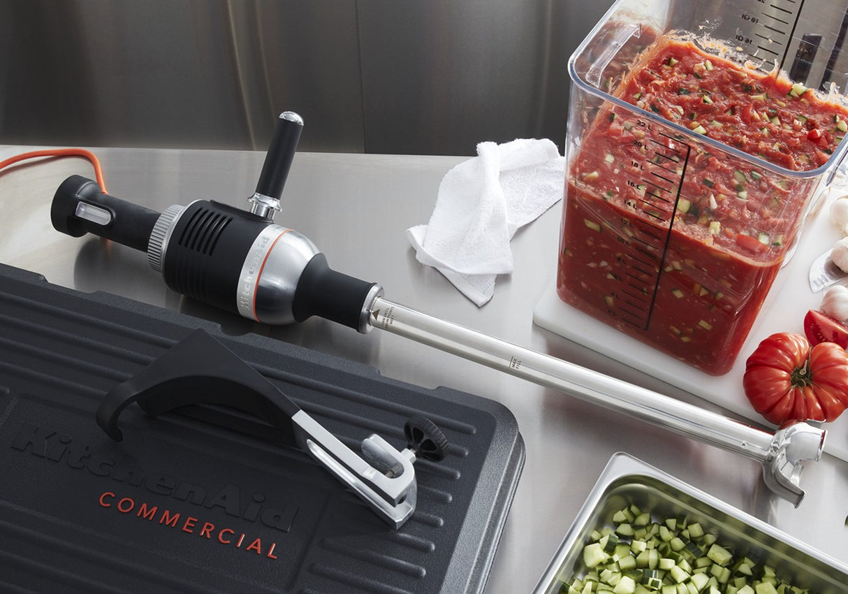 A heavy duty immersion blender lies on a stainless steel counter near a large container of diced tomatoes and cucumbers. A folded white towel, a black KitchenAid case, and chopped vegetables in a clear container are also visible.