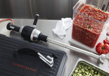 A heavy duty immersion blender lies on a stainless steel counter near a large container of diced tomatoes and cucumbers. A folded white towel, a black KitchenAid case, and chopped vegetables in a clear container are also visible.