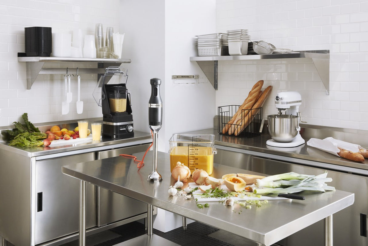 A modern commercial kitchen with stainless steel counters and shelves. Fresh vegetables, chopped herbs, and soup in a measuring container sit on the table. A KitchenAid Commercial Blender with 60 oz. capacity is neatly arranged among baguettes, dishes, and utensils.