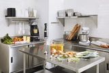 A stainless steel commercial kitchen features shelves with plates, glasses, utensils, and baguettes. Nearby, a KitchenAid commercial immersion blender with a 12 stainless steel blending arm sits beside prepared vegetables and a container of soup. White tile walls complete the clean look.