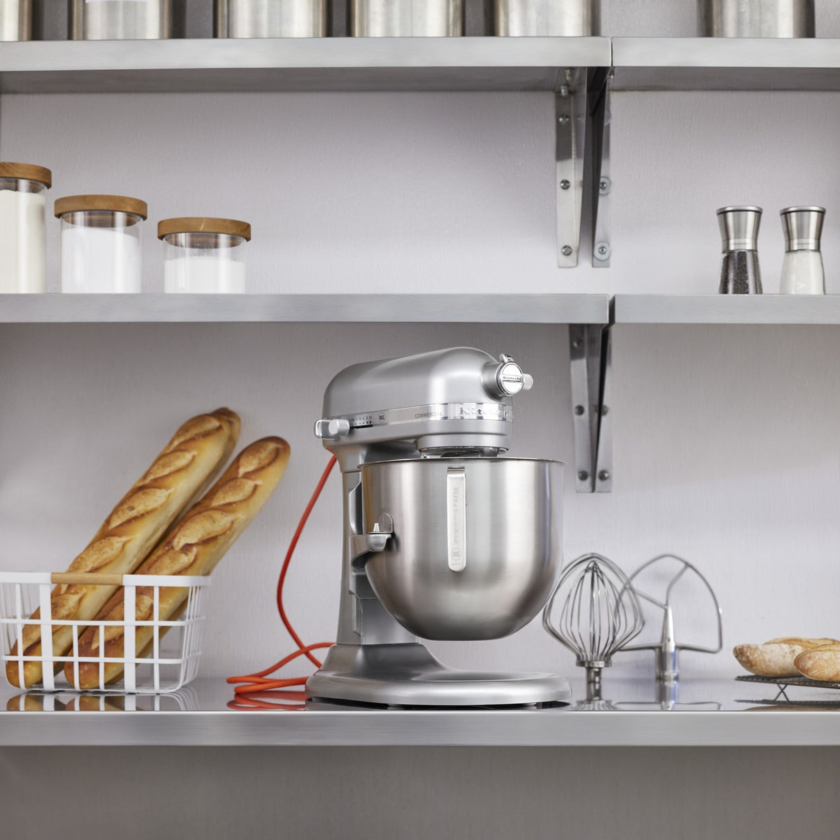 A silver KitchenAid Commercial Stand Mixer with an 8 quart bowl and attachments sits on a kitchen counter. Nearby are a wire basket with two baguettes, jars of ingredients, and pastries. Metal shelves with containers hang above against a white wall.