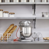 A KitchenAid Commercial Stand Mixer with an 8 quart bowl sits on a kitchen counter beside mixing attachments and a wire basket holding two baguettes. Above, shelves hold jars, canisters, and salt and pepper shakers in this clean, minimalist kitchen.