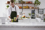 A man in a white shirt and black apron places two green smoothies on a marble counter in a bright juice bar, with fresh fruit, juices, and an Enclosure Blender with 60 oz. capacity on shelves behind him. A chalkboard menu hangs on a tiled wall.