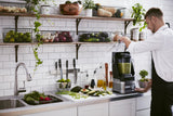 A man in an apron stands in a modern kitchen, blending green smoothie ingredients with a KitchenAid Commercial Blender. The countertop holds fresh vegetables and utensils, while open shelves display produce and jars above a white tile backsplash.