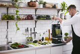 A man in a white shirt and black apron uses a KitchenAid Commercial Blender with a 60 oz. capacity to prepare a green smoothie in a modern kitchen with white tile walls, open shelves of produce, and fresh fruits and vegetables on the counter.