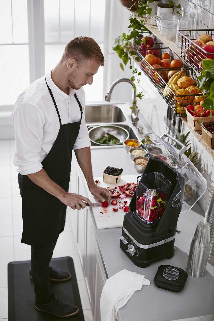 A man in a white shirt and black apron stands in a bright kitchen, slicing strawberries beside a KitchenAid Commercial Blender with 60 oz. capacity. Fresh produce and bowls of ingredients line the counter, while shelves above hold baskets of colorful fruit.