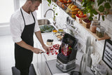 A man in a black apron slices fruit on a cutting board in a modern kitchen. He prepares ingredients for his BPA free blender, which contains chopped fruit. The counter holds bowls of fresh produce, with plants and kitchenware visible above.