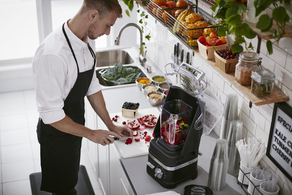 A man in a black apron slices strawberries on a white cutting board beside a KitchenAid Commercial Blender filled with fruit. The bright, organized kitchen features bowls of greens, glasses, plants, and shelves stocked with produce.