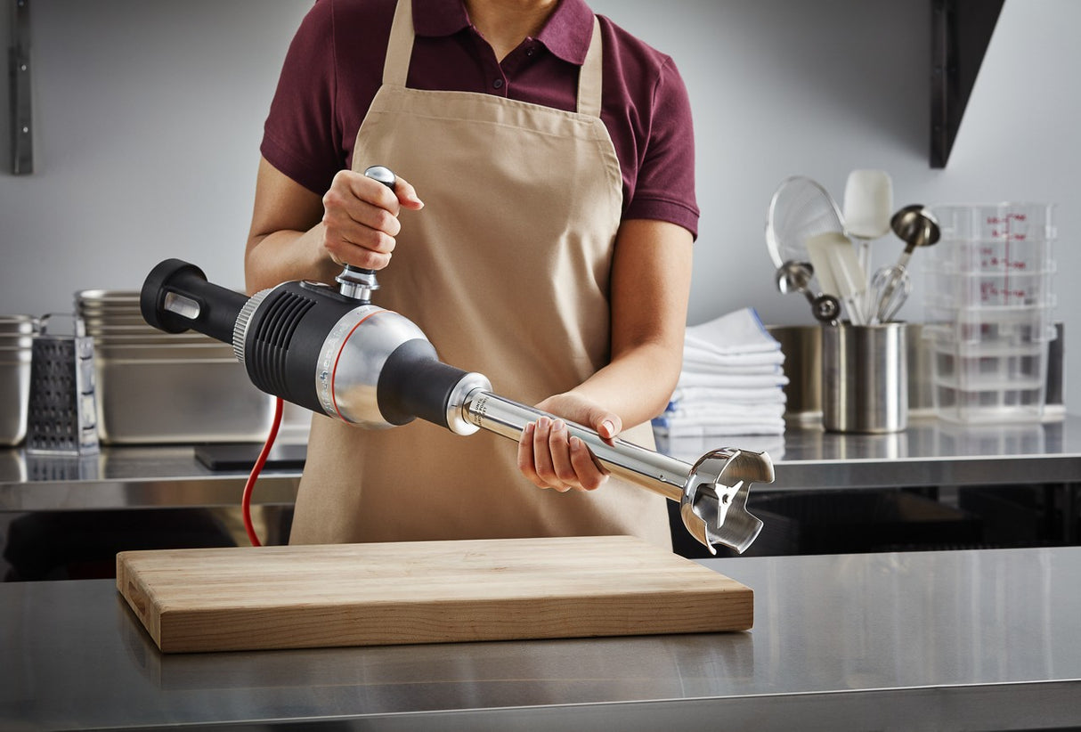 A person in a maroon shirt and beige apron holds a heavy duty immersion blender over a wooden cutting board in a stainless steel kitchen, with utensils, white towels, and containers visible in the background.