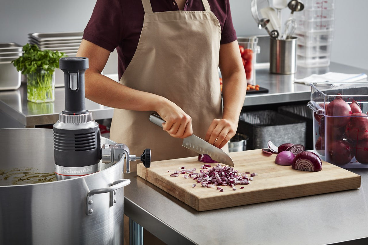 A person in a beige apron chops red onions on a wooden cutting board in a commercial kitchen. Chopped and halved onions sit nearby, with a large pot, fresh herbs, and a heavy duty immersion blender in the background.