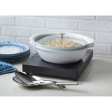 A white ceramic pot with a glass lid sits on a low profile induction cooktop. The pot holds a chunky vegetable stew. Two metal serving spoons rest nearby, with blue plates and a folded blue cloth stacked on the left against a white brick background.