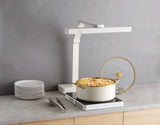 A white pot with gold handles, filled with pasta and vegetables, sits on a sleek low profile induction cooktop. The lid stands upright on a holder. Nearby are stacked plates and utensils, beneath a modern kitchen lamp on the light gray countertop.