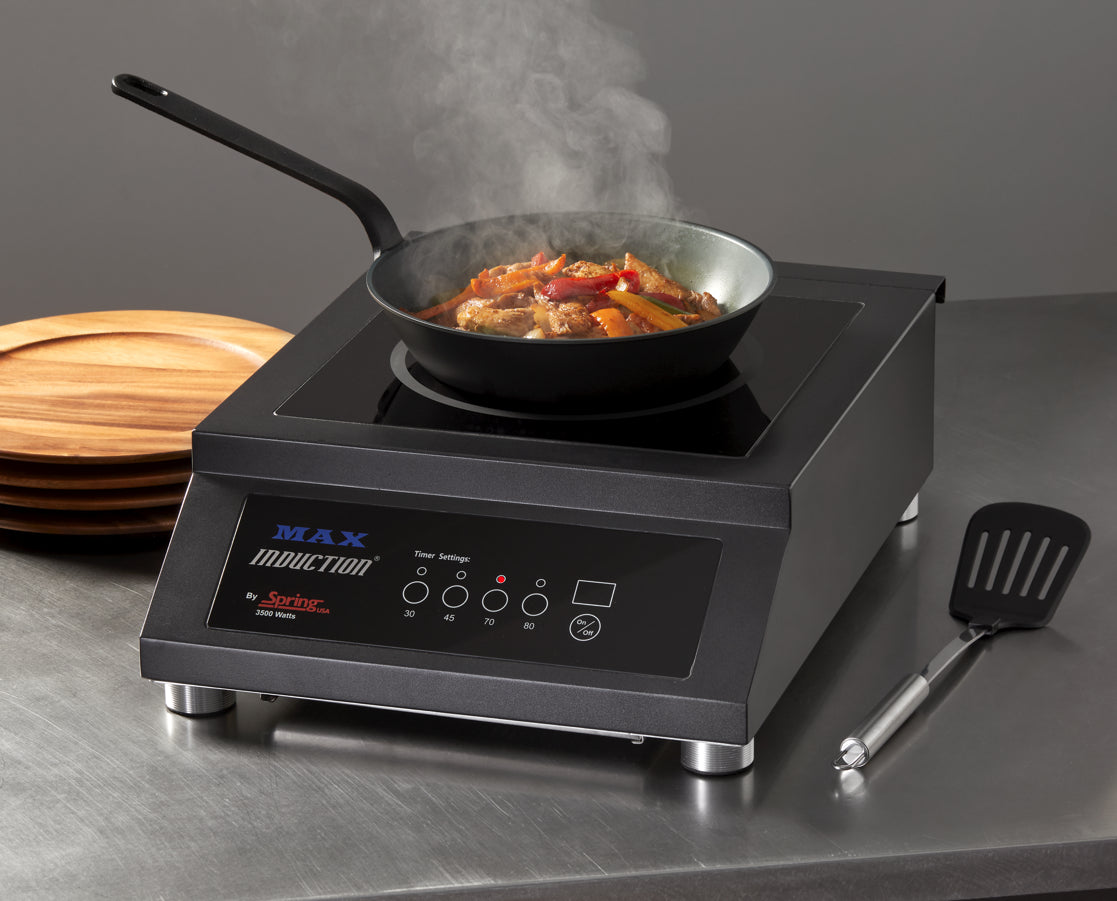 A black countertop induction cooktop with a digital control panel sits on a stainless steel counter. A frying pan with steaming stir-fried vegetables and meat cooks on top. Nearby are stacked wooden plates and a metal spatula with a black handle.
