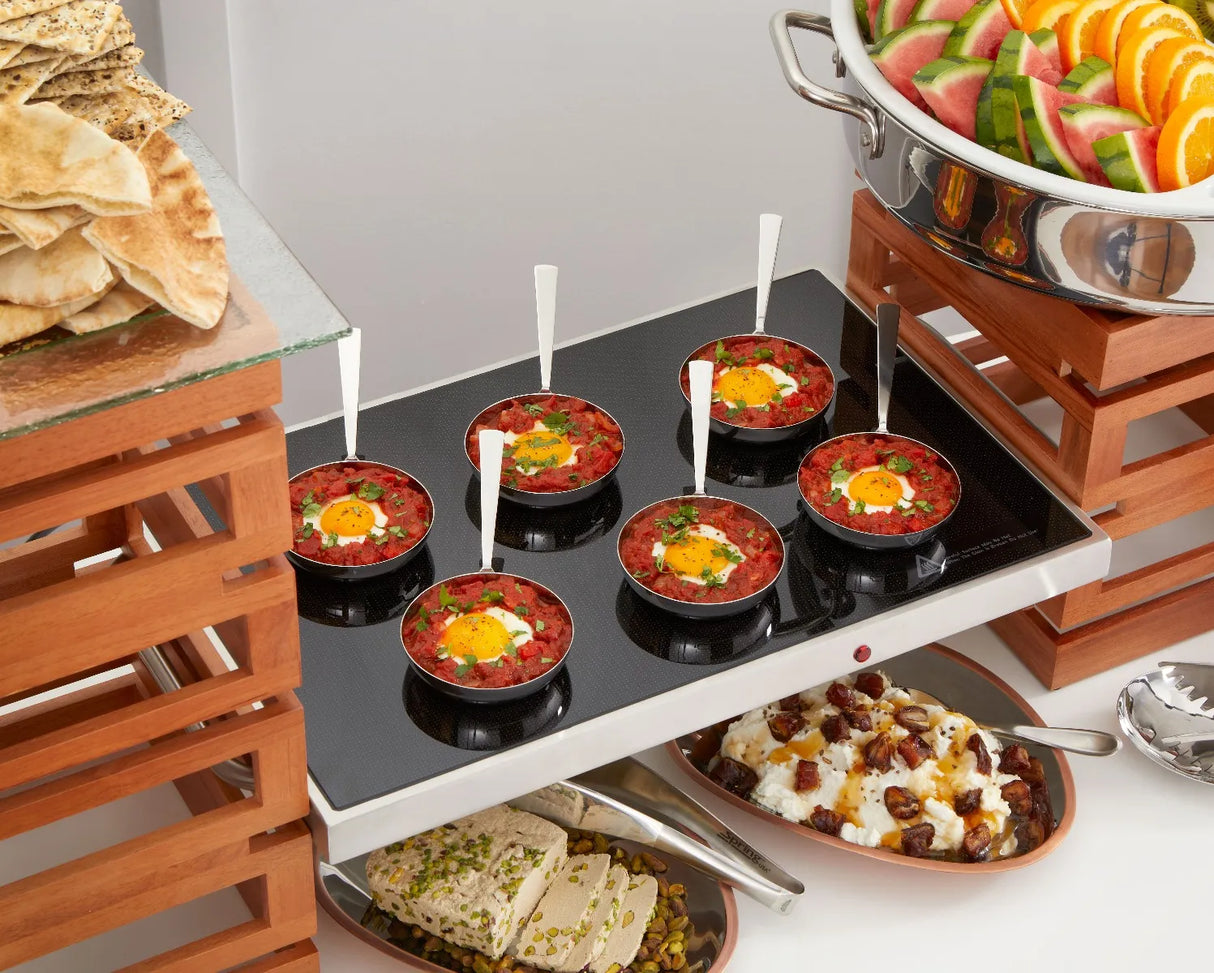 Small pans of shakshuka with eggs are displayed on a warming tray with white-handled spoons. Surrounding trays hold sliced pita bread, a fruit bowl with watermelon and orange, herbed white cheese, and yogurt topped with dates.
