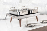 A stainless steel buffet warmer with a sturdy stainless steel body and two rectangular trays sits on a white marble countertop. Wooden legs support the warmer, while two serving spoons rest nearby against white marble tile.