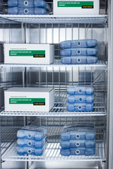 Boxes labeled “Varicella Virus Vaccine Live Varivax” and several blue ice packs are arranged on metal wire refrigerator shelves inside a commercial reach-in freezer. The environment looks sterile, organized, and cold.