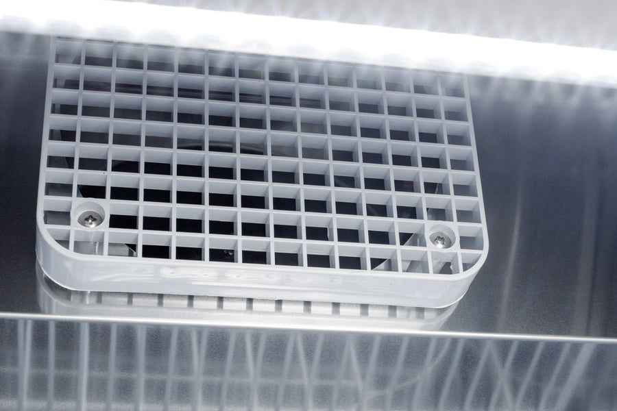 Close-up of a metallic vent on a pharma-lab all-freezer with a white plastic grid cover, secured by two screws. Bright LED lights above cast reflections on the shiny metal surface, creating parallel light streaks across the image.