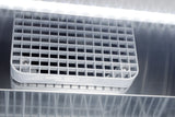 Close-up of a metallic vent on a pharma-lab all-freezer with a white plastic grid cover, secured by two screws. Bright LED lights above cast reflections on the shiny metal surface, creating parallel light streaks across the image.