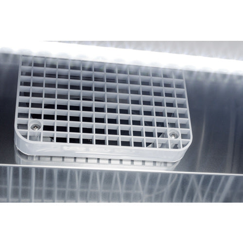 A close-up of a white plastic vent with a grid pattern secured by two screws on the reflective metallic surface of a commercial refrigerator, illuminated by bright white light from above, creating light streaks and reflections.